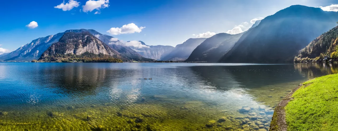 Panoramablick auf einen klaren See mit einer glatten Wasseroberfläche, umgeben von hohen, bewaldeten Bergen. Am sonnenbeschienenen Ufer sieht man klares, steiniges Wasser und eine Grasfläche. Ein paar Enten schwimmen auf dem See.