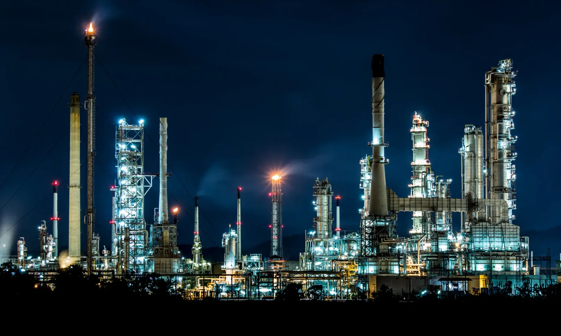 Nächtliche Ansicht einer großen Industrieanlage, die von zahlreichen Lichtern erhellt wird. Die Anlage besteht aus mehreren hohen Türmen und Schornsteinen, von denen einer eine große Flamme ausstößt. Die Beleuchtung hebt die metallischen Strukturen und Rohrleitungen der Anlage hervor und schafft ein beeindruckendes und intensives Bild. Die dunkle Nacht bildet einen starken Kontrast zu den hell erleuchteten Elementen der Anlage, wodurch die industrielle Tätigkeit und Komplexität betont wird.